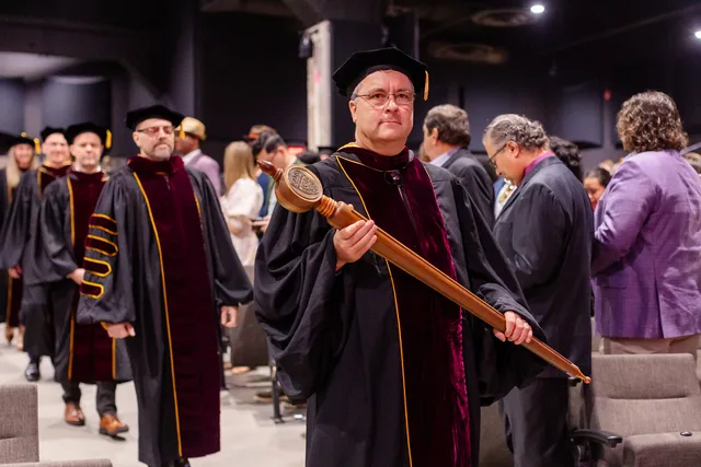 Robed faculty member carrying the ceremonial mace, procession behind him.