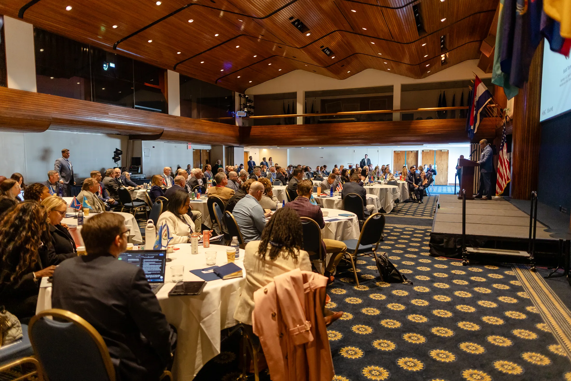 Wide view of a packed hall of attendees at round tables looking toward a lit stage under a wood-paneled ceiling.