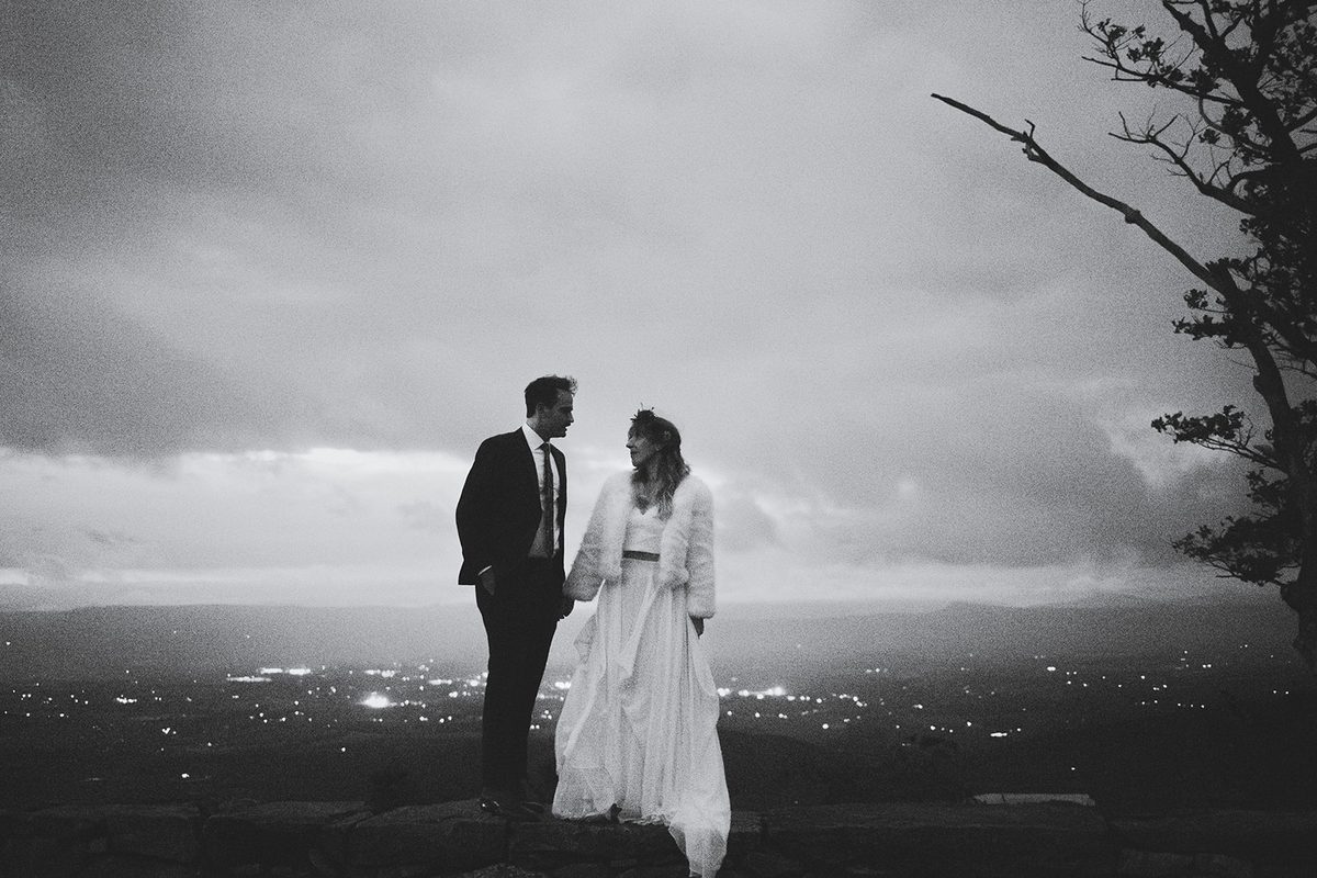 A couple facing each other on a summit at night with the lights of the valley behind them