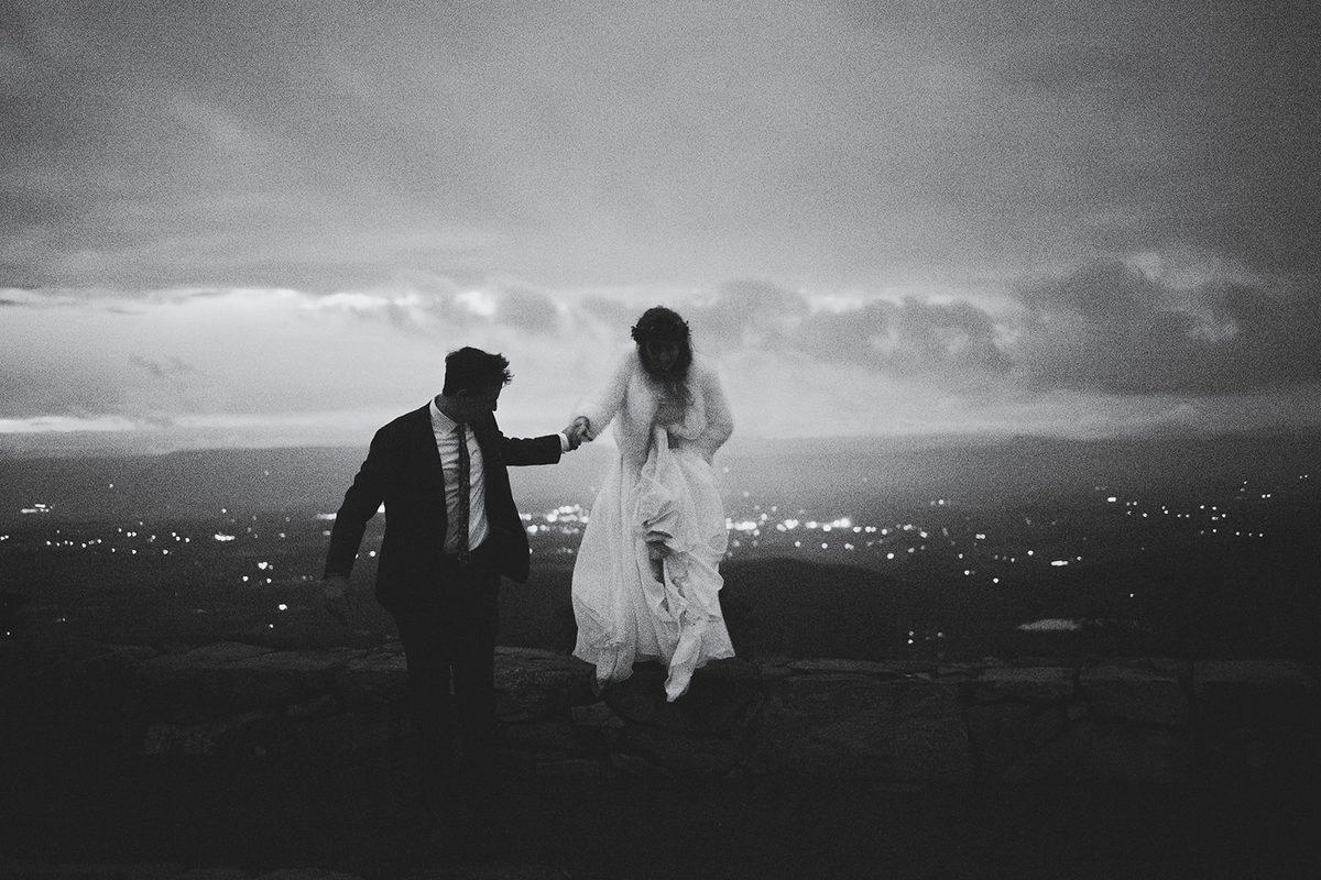 A groom leading his bride along a dark stone overlook, her skirt trailing behind her