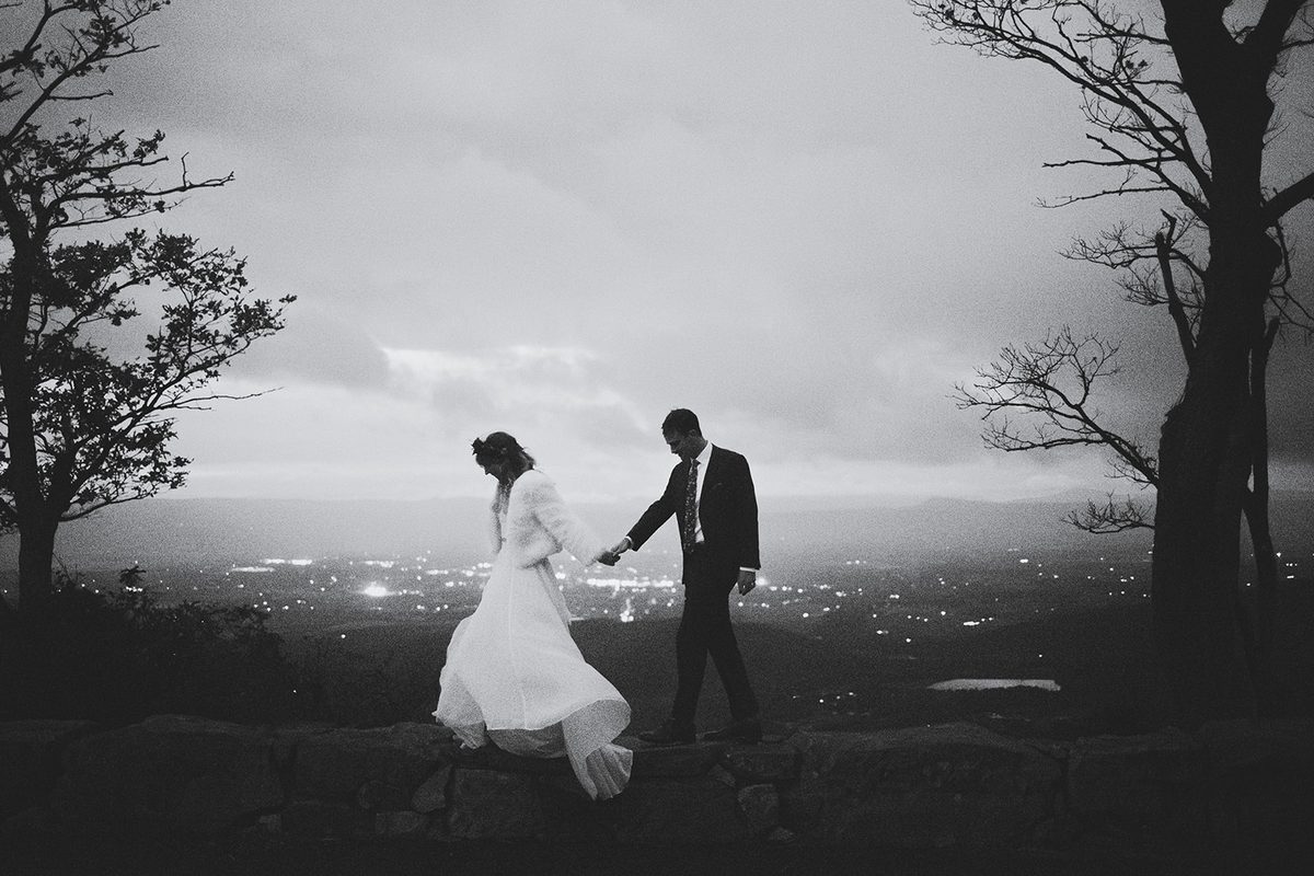 A couple walking a dark ridge line framed by a bare tree, valley lights glittering below