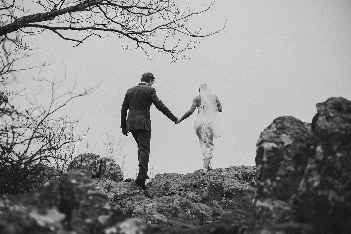 A groom leading his bride by the hand across a bare rocky ridge under a pale sky