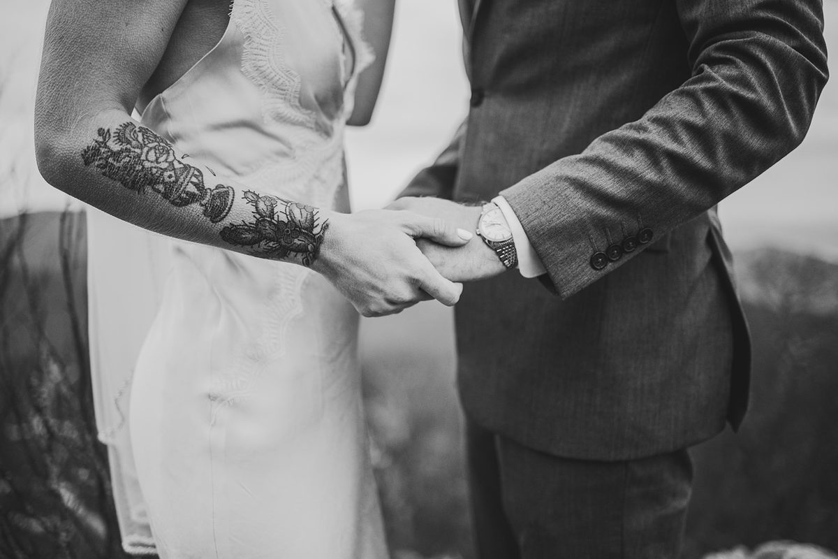 A close frame of a couple's clasped hands at their sides, her tattooed forearm against his suit