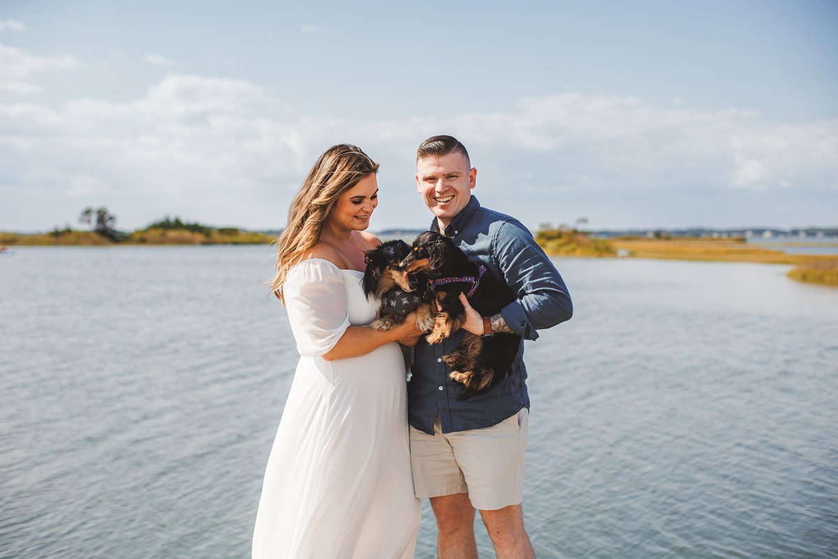A couple laughing on a dock while holding their long-haired dachshund between them