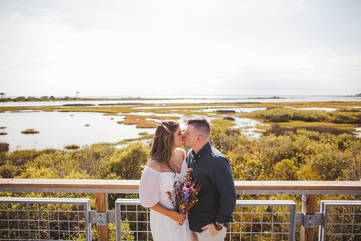 A couple kissing at a railing above a wide green salt marsh under an open sky