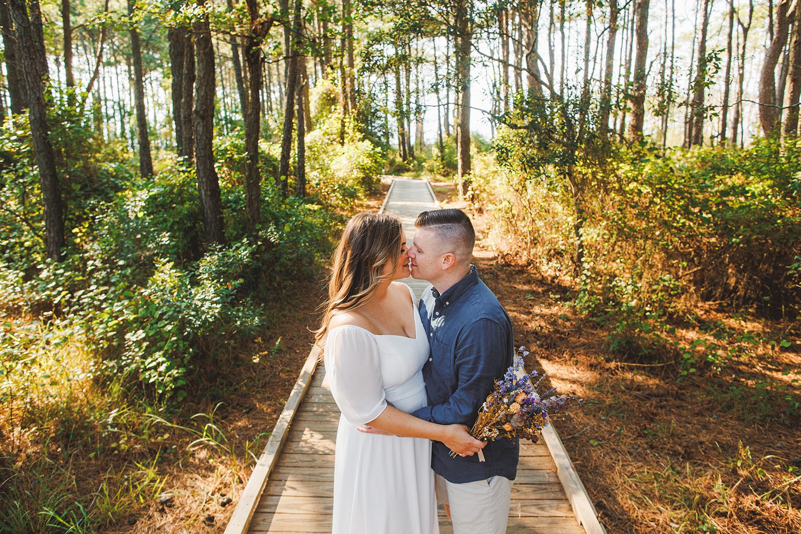 A couple kissing on a wooden boardwalk running through tall coastal pines