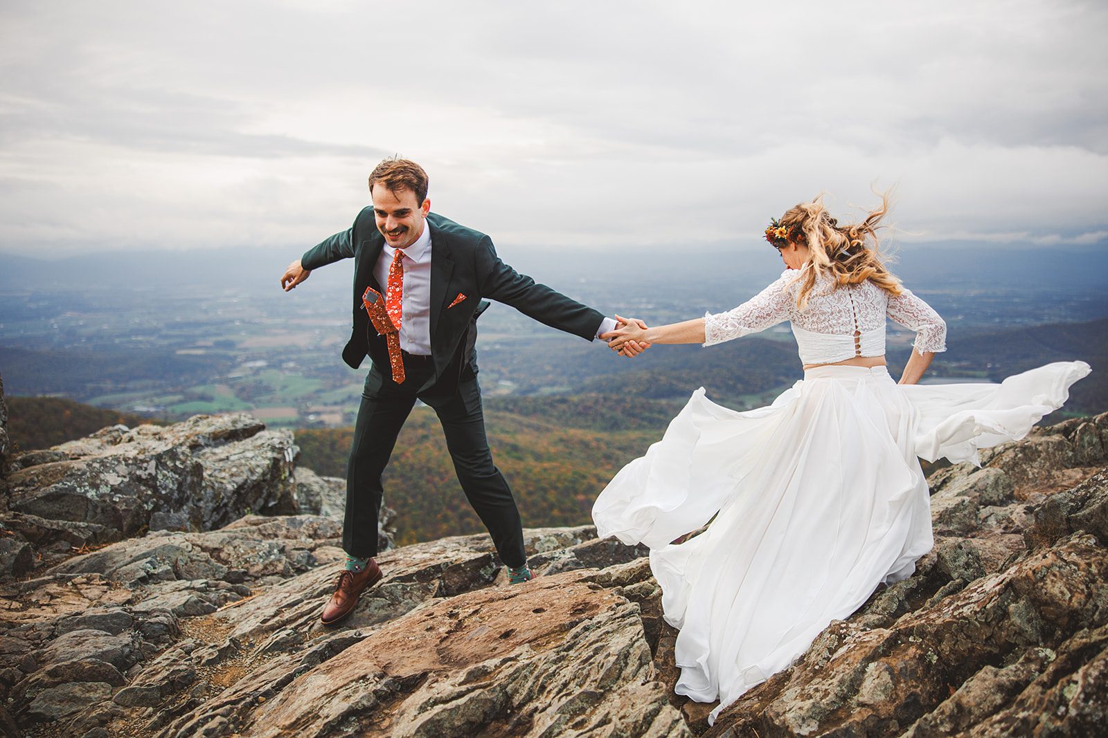 A couple on a windy Shenandoah summit, her skirt sweeping out behind her as he holds her hand on the rock