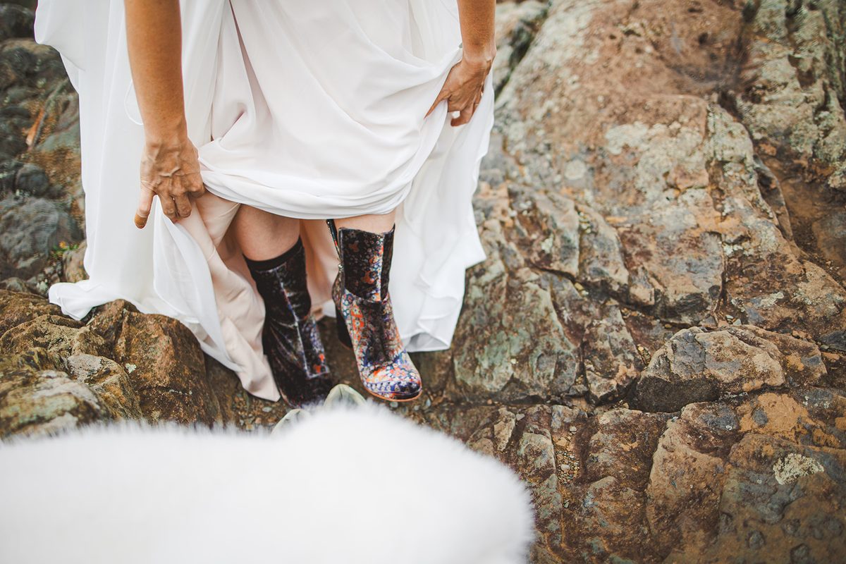 A bride lifting her gown to show patterned hiking boots as she climbs a rock face