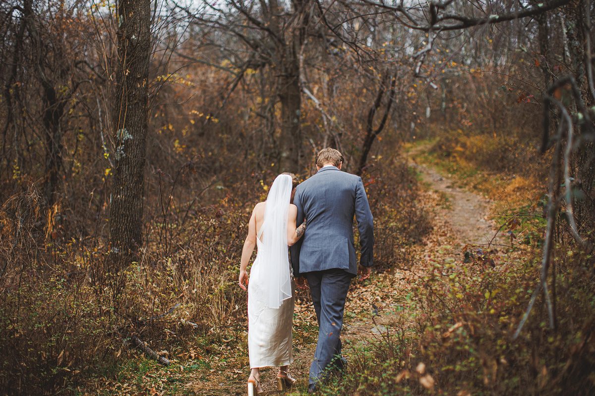 A couple walking away up a narrow trail through bare autumn woods, her veil catching the light
