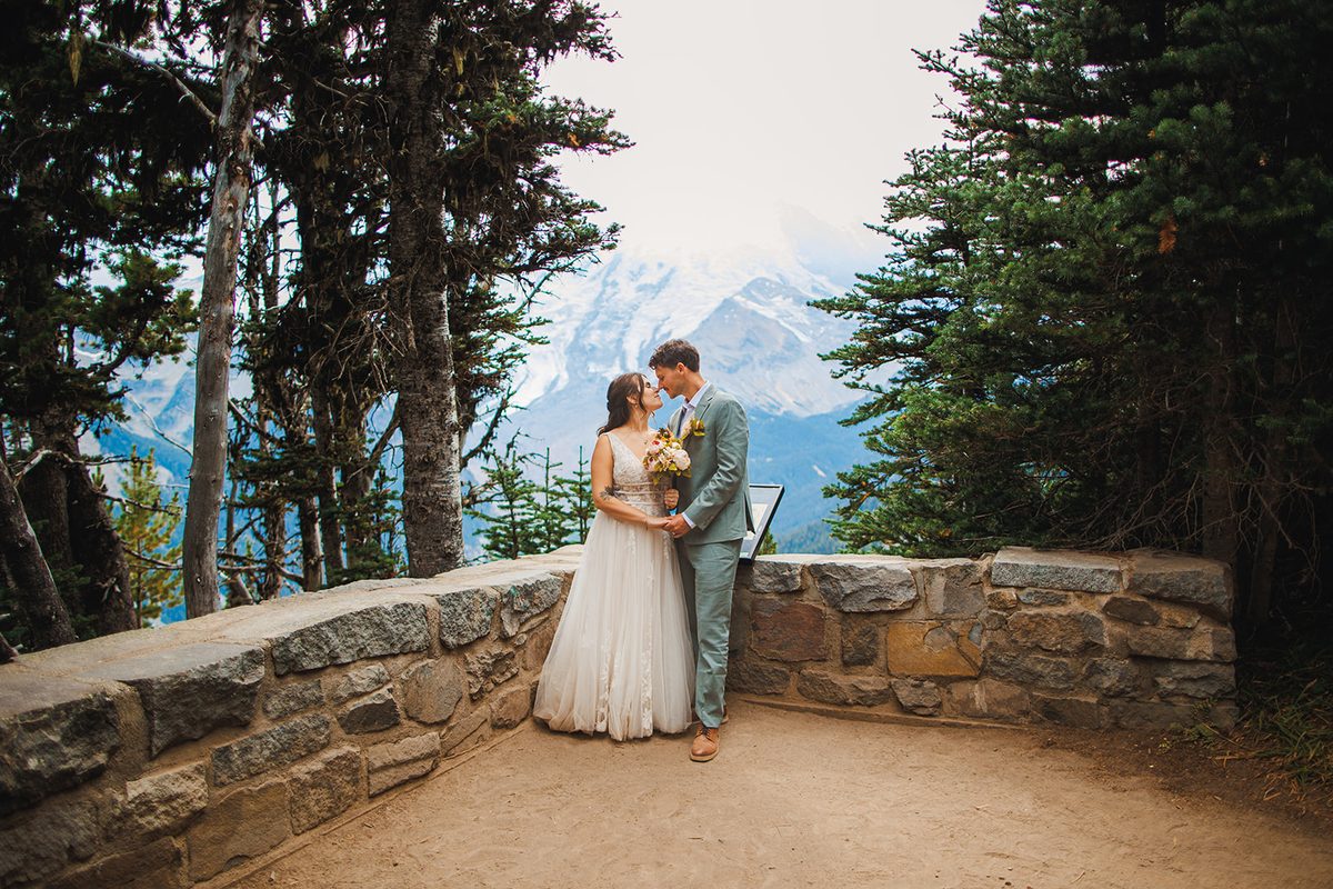 A couple saying their vows at a stone overlook framed by firs, a glacier-covered peak between them