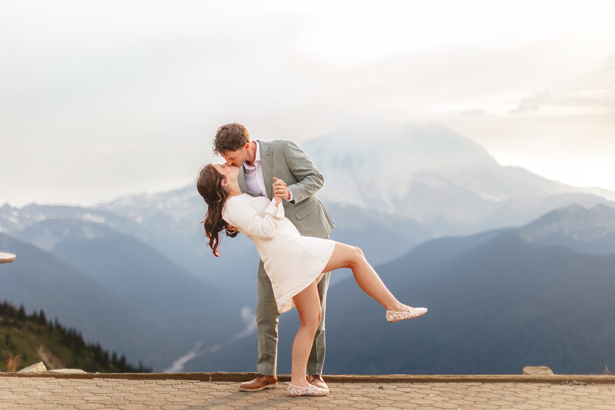 A groom lifting and kissing his bride on an overlook with a hazy mountain range falling away behind them