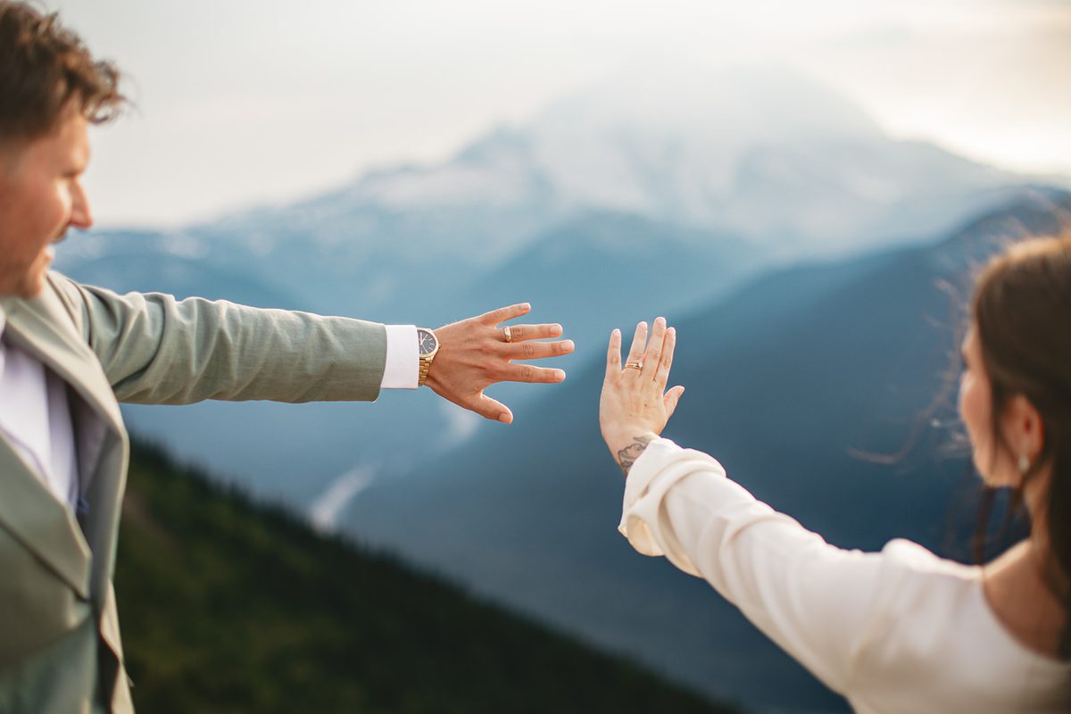A couple reaching their hands toward each other, both rings visible, mountains blurred behind
