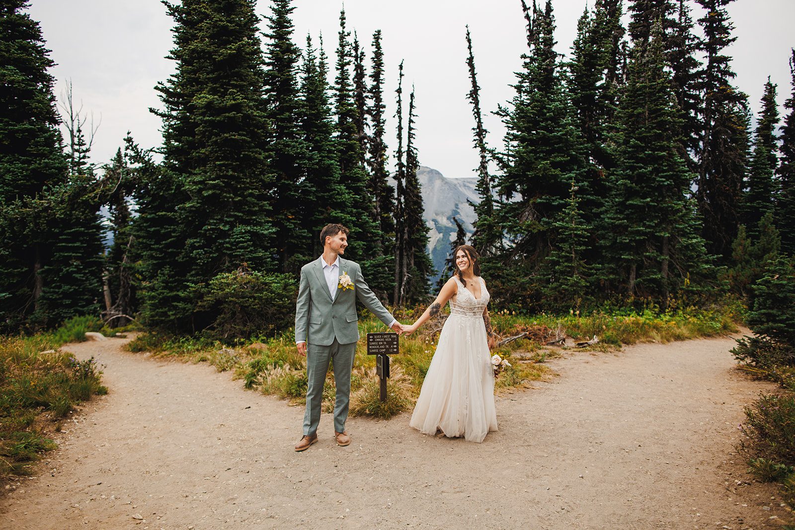 A couple holding hands at a mountain trail junction framed by tall firs with a snow-covered peak behind