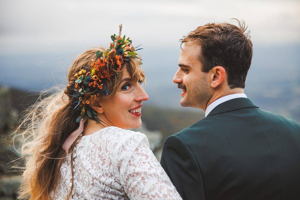 A bride in an autumn flower crown glancing back over her shoulder, hair lifting in the wind, the groom looking at her