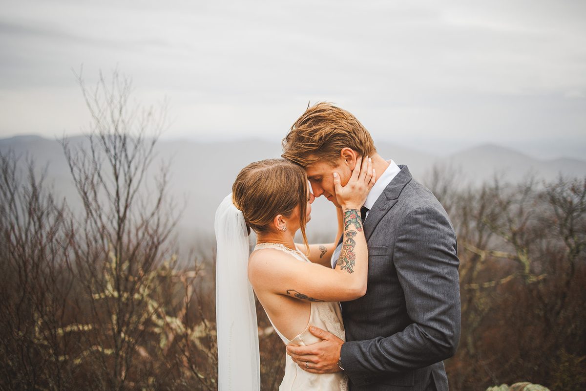A couple holding each other's faces on a ridge with soft blue mountain layers behind them