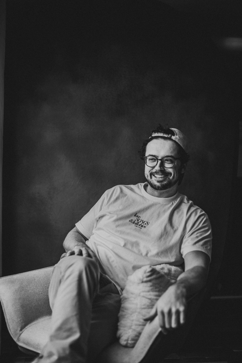 Aaron May sitting back in a chair with an easy smile, in a backwards cap and glasses, photographed in black and white