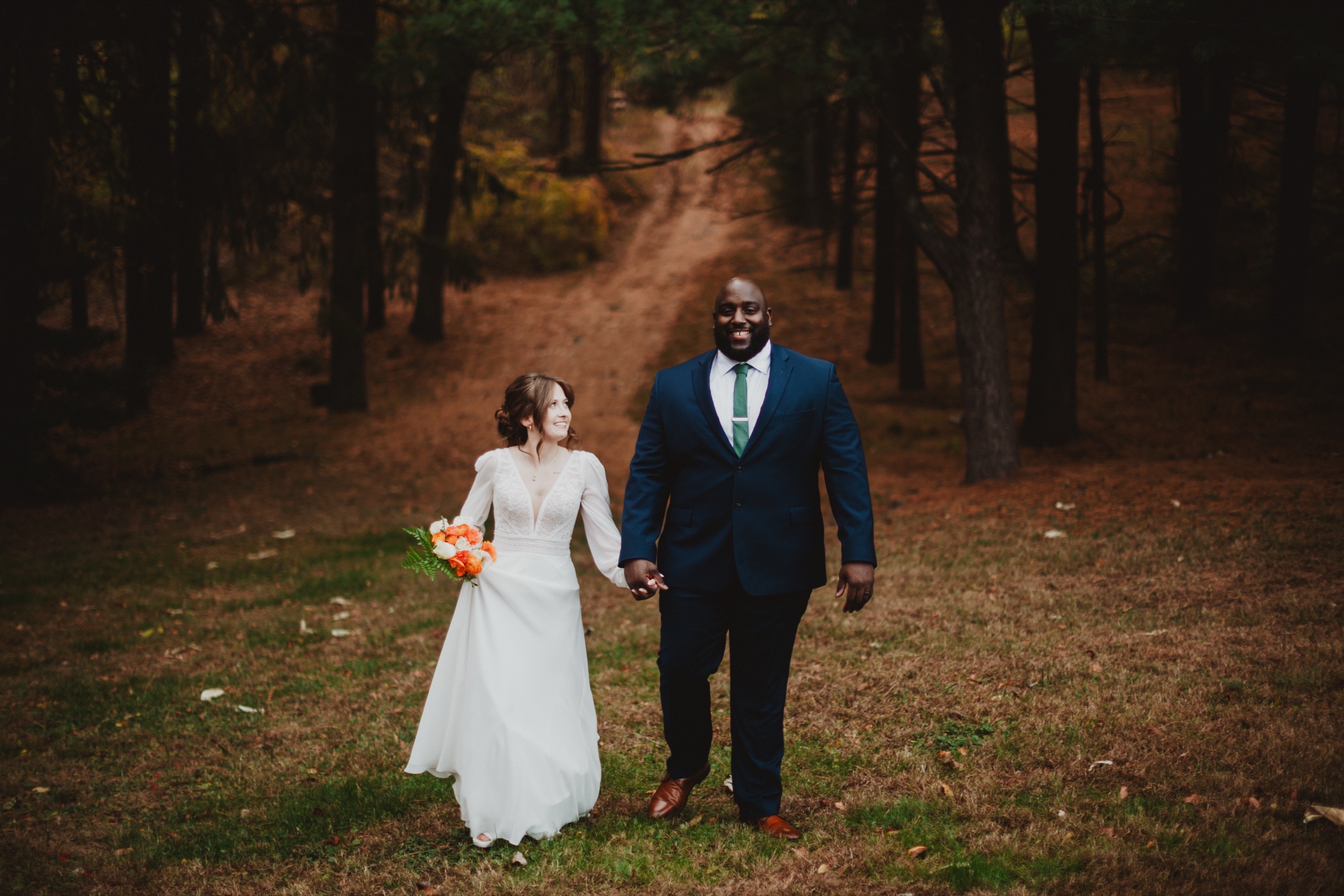 Delaney and Dante walking hand in hand through the pines after their ceremony, 2025