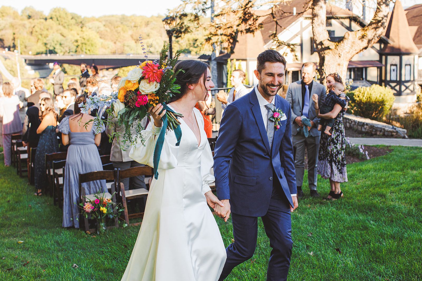 The groom grins as he and the bride walk hand in hand across the lawn, her bouquet lifted over her shoulder, guests standing among the ceremony chairs behind them.