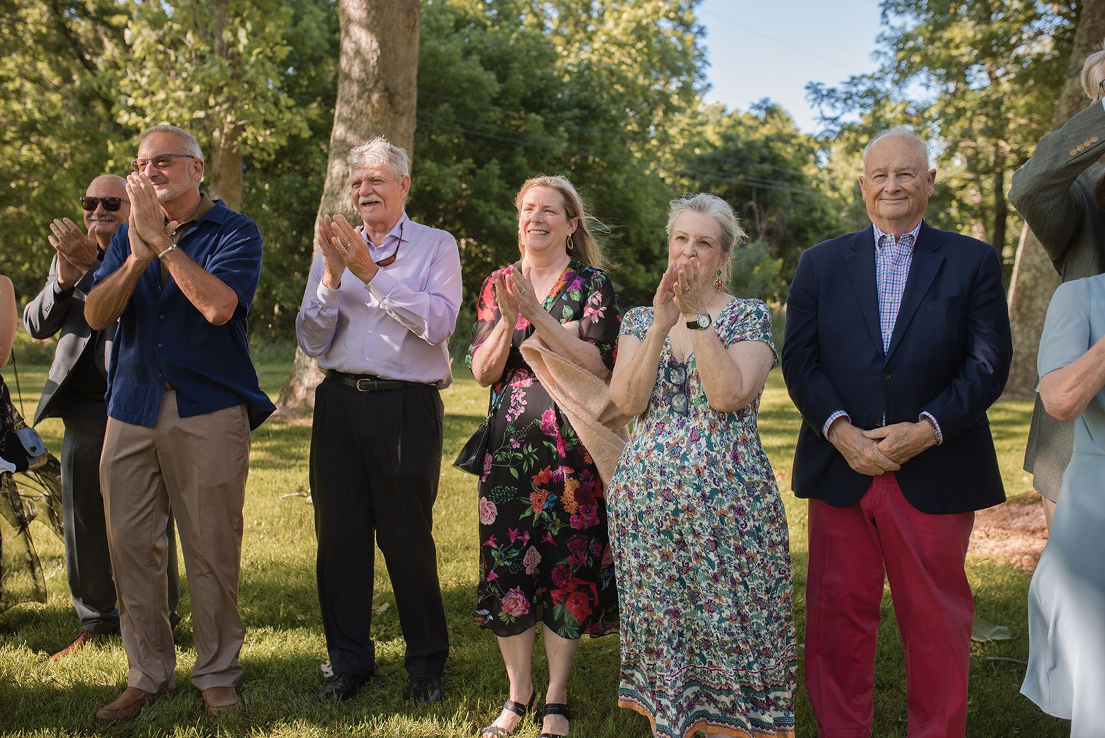 A row of guests stands clapping on the grass with trees behind them, several of them smiling.