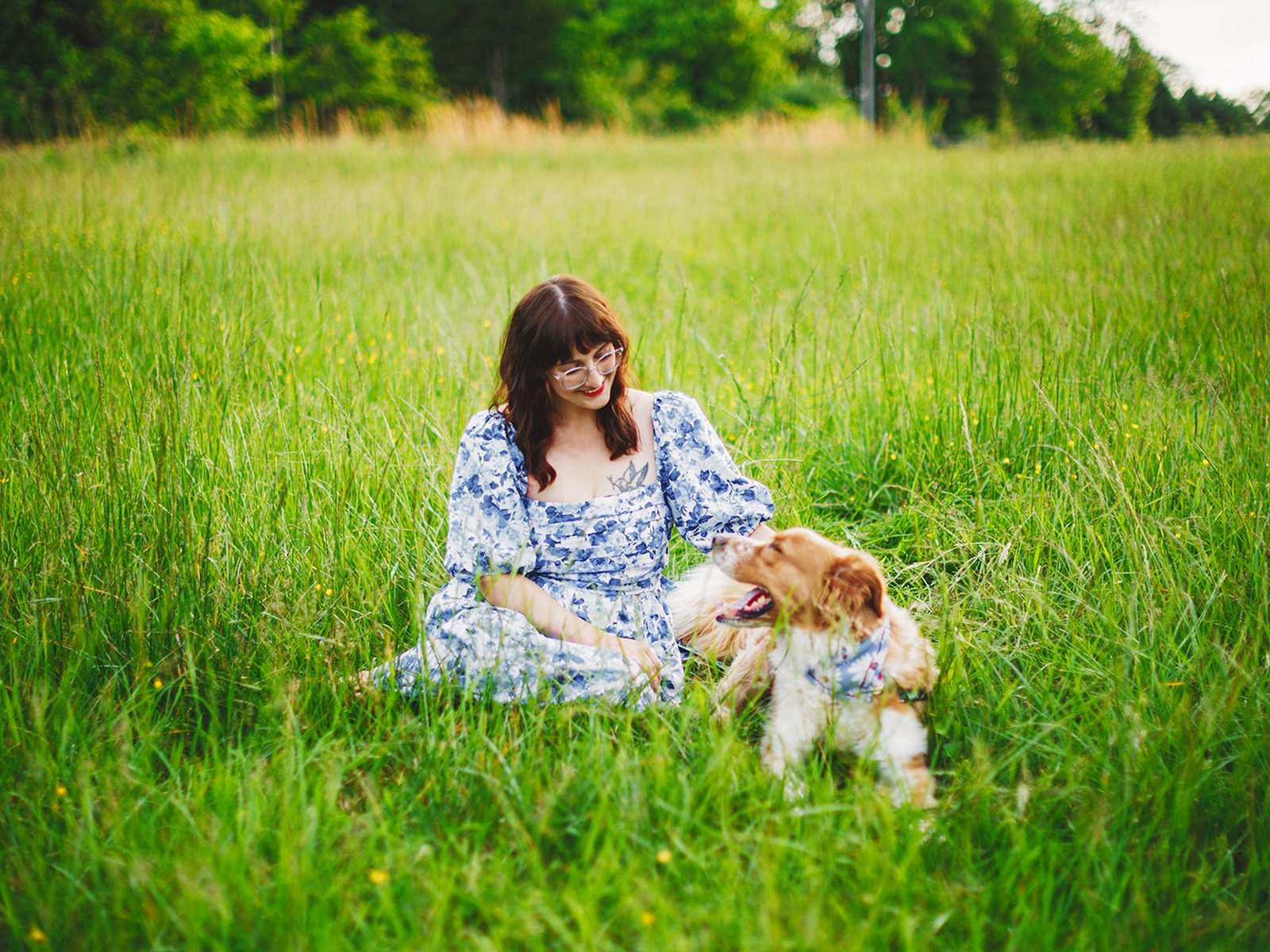 A woman sitting down in a green meadow with her dog lying in the grass beside her, both relaxed