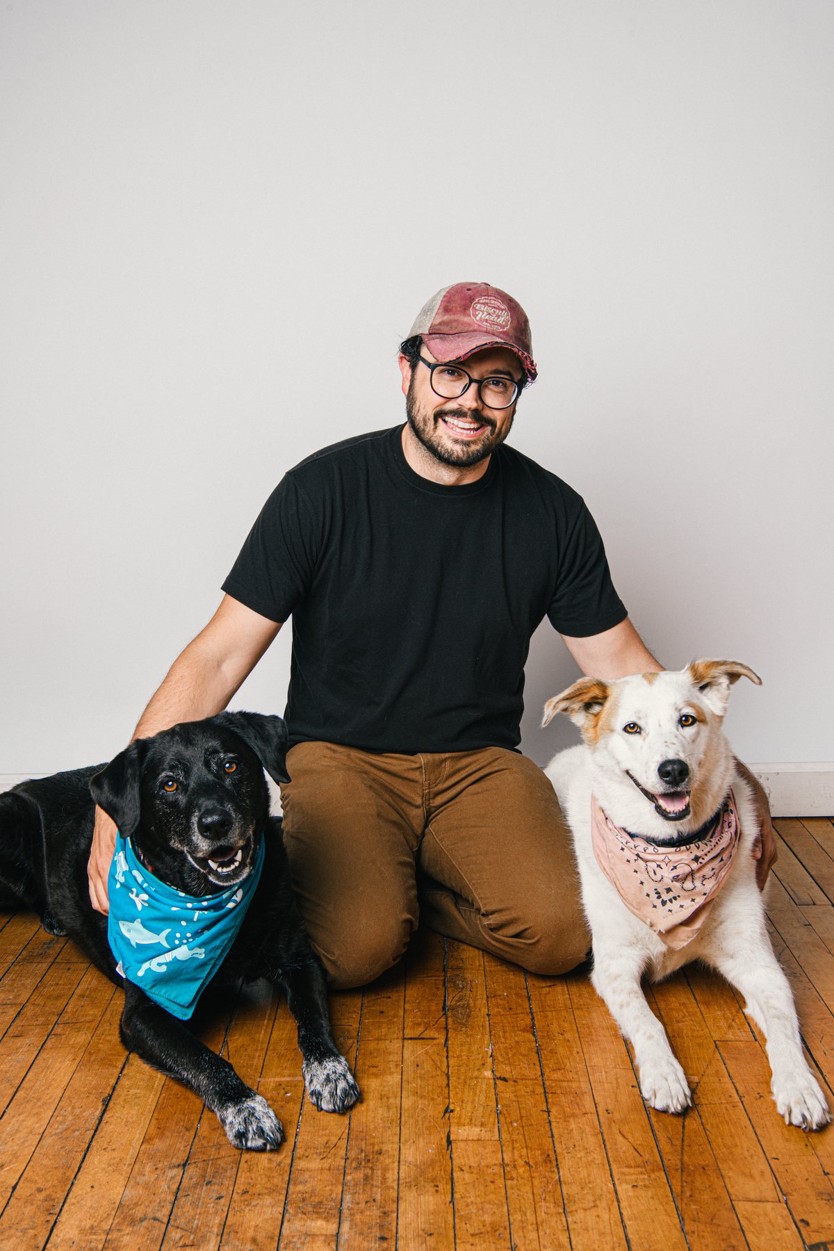 Aaron kneeling on a wood floor between his two dogs: Cal, a black lab in a blue shark bandana, and Sugar, a white dog in a pink bandana, both lying down and smiling