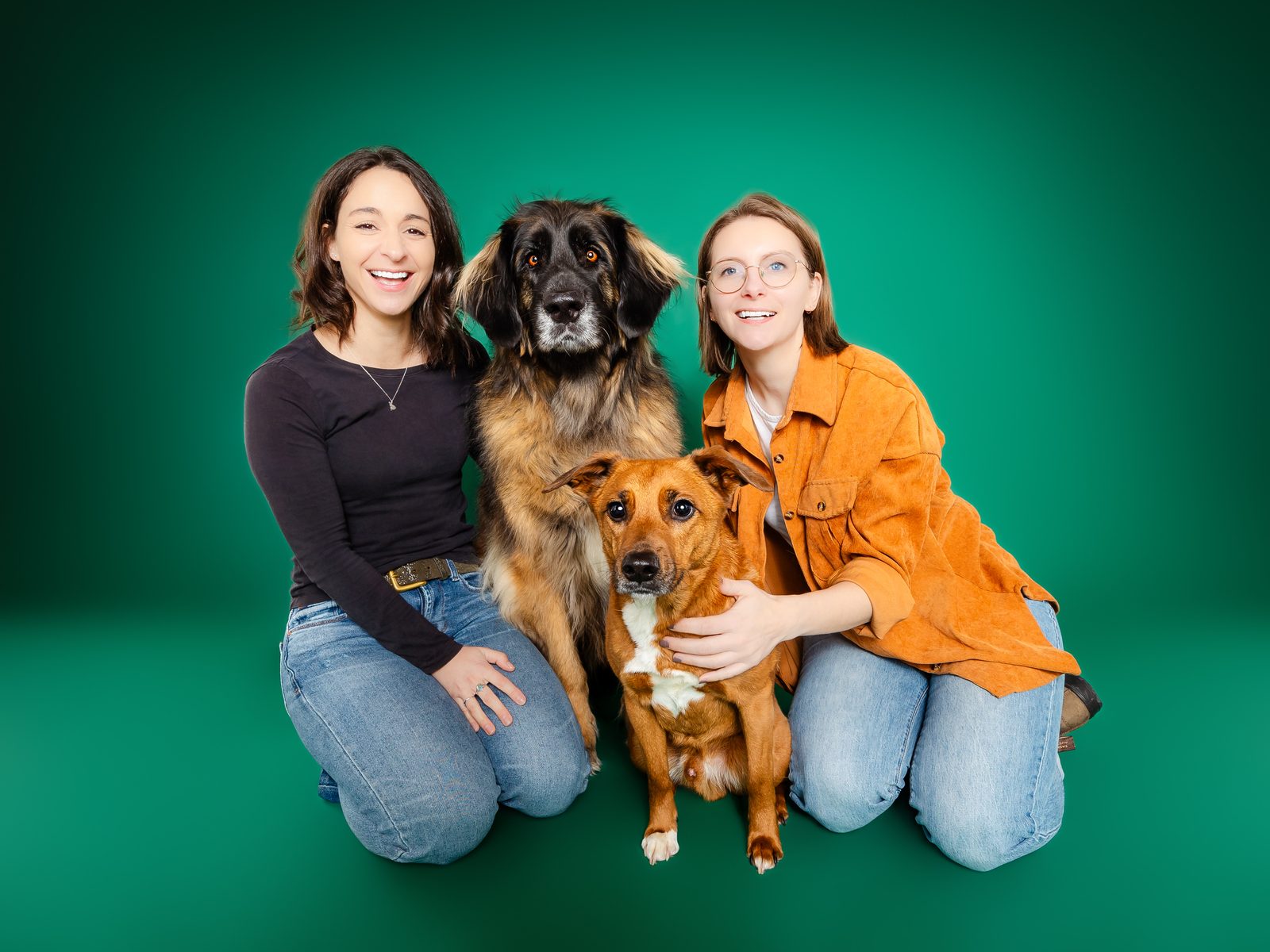 Two people kneeling on a kelly green backdrop with their two dogs between them, everyone looking at the camera