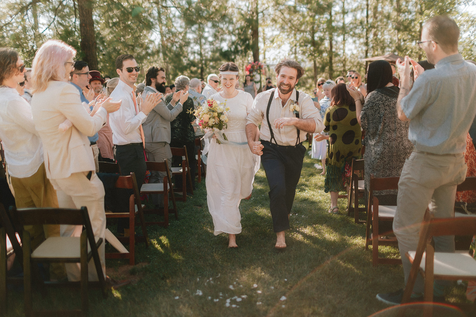A barefoot couple running back up the aisle through applauding guests in golden woods