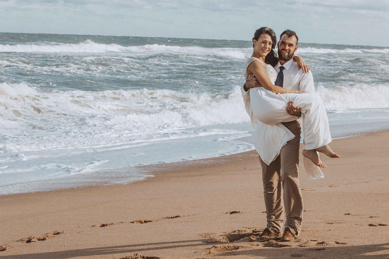 Groom carrying the bride along the surf line on their wedding day, both laughing