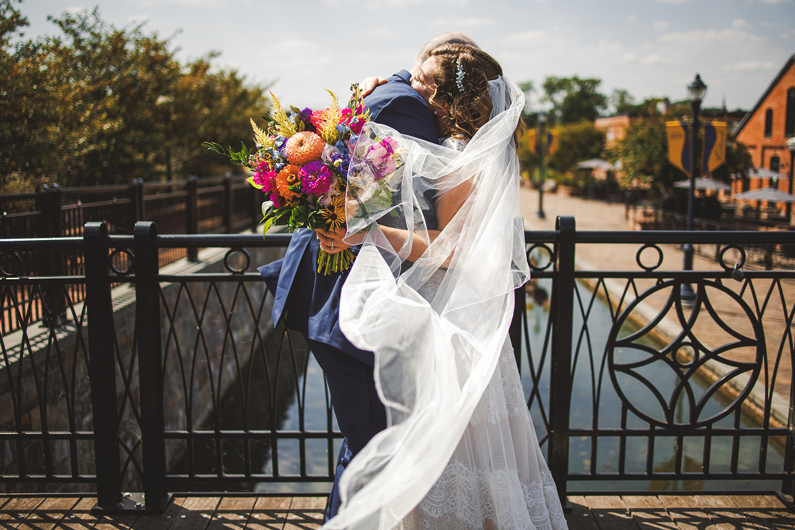 Alex and Phil embracing on the Carroll Creek footbridge in downtown Frederick, the veil sweeping across a bright wildflower bouquet
