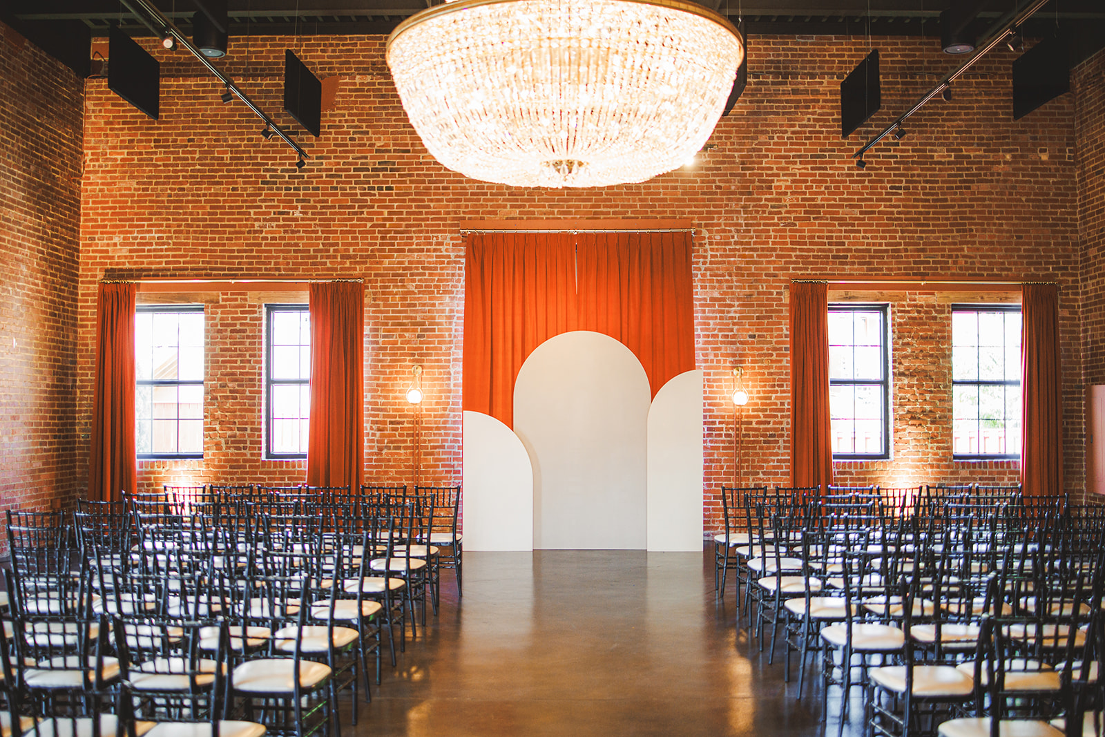 The ceremony room at Union Mills Public House: brick walls, a crystal chandelier, and a white arch against an orange curtain