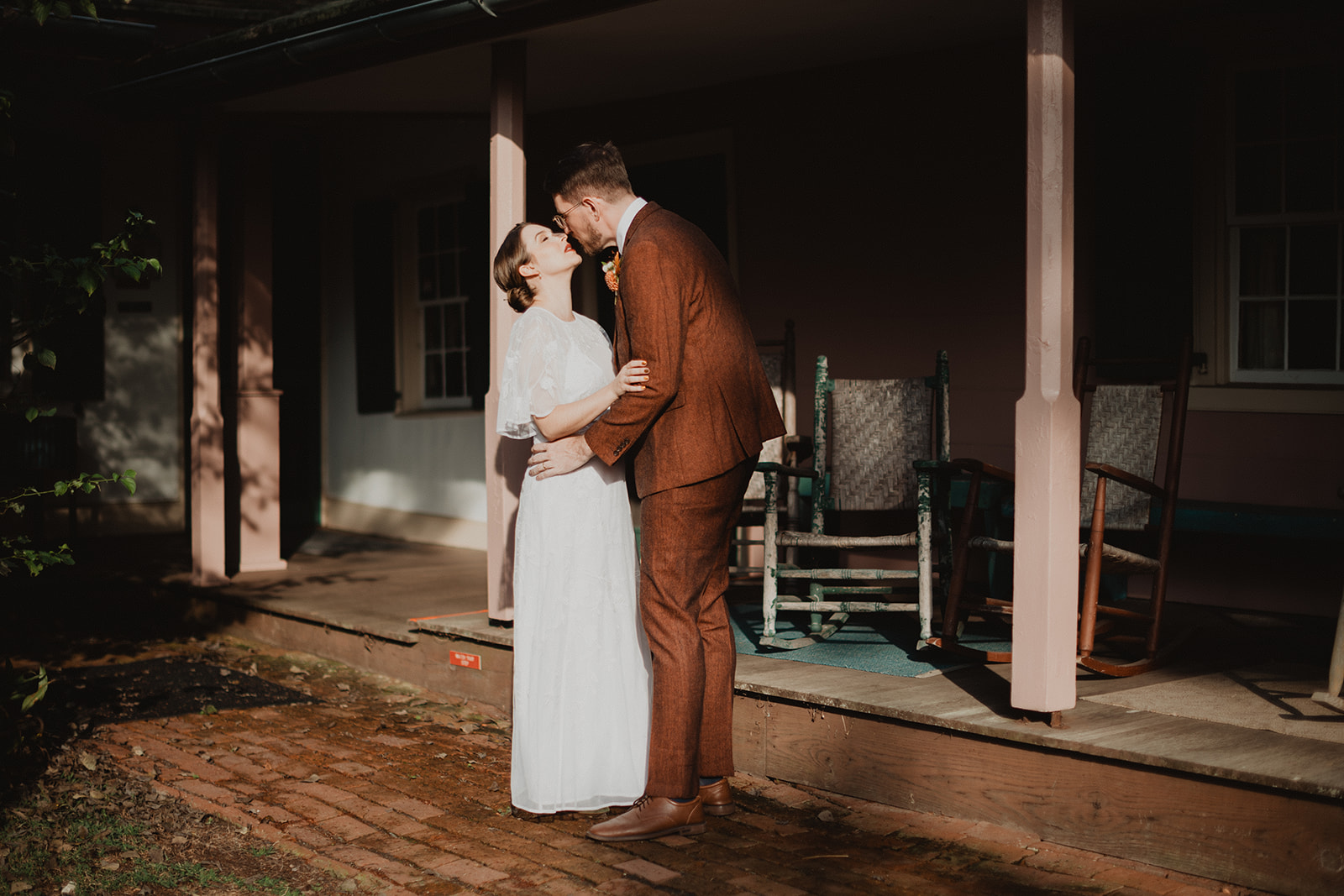 Josh kissing Alison on the farmhouse porch at Union Mills Homestead, the two of them carved out of hard afternoon light