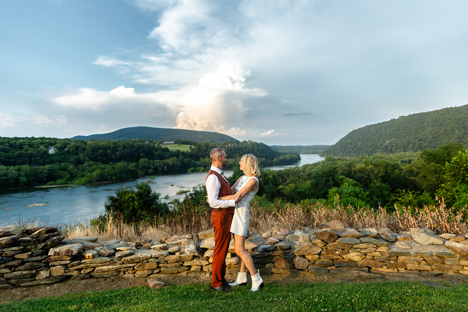 Annie and Thomas embracing at the stone wall at Harpers Ferry Brewing, the Potomac winding through the valley below under a towering evening cloud