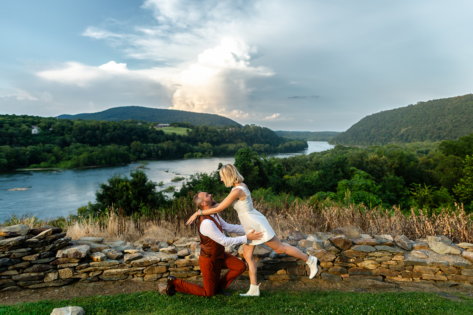 Thomas on one knee at the overlook wall as Annie leans into his arms laughing, the river valley spread out behind them