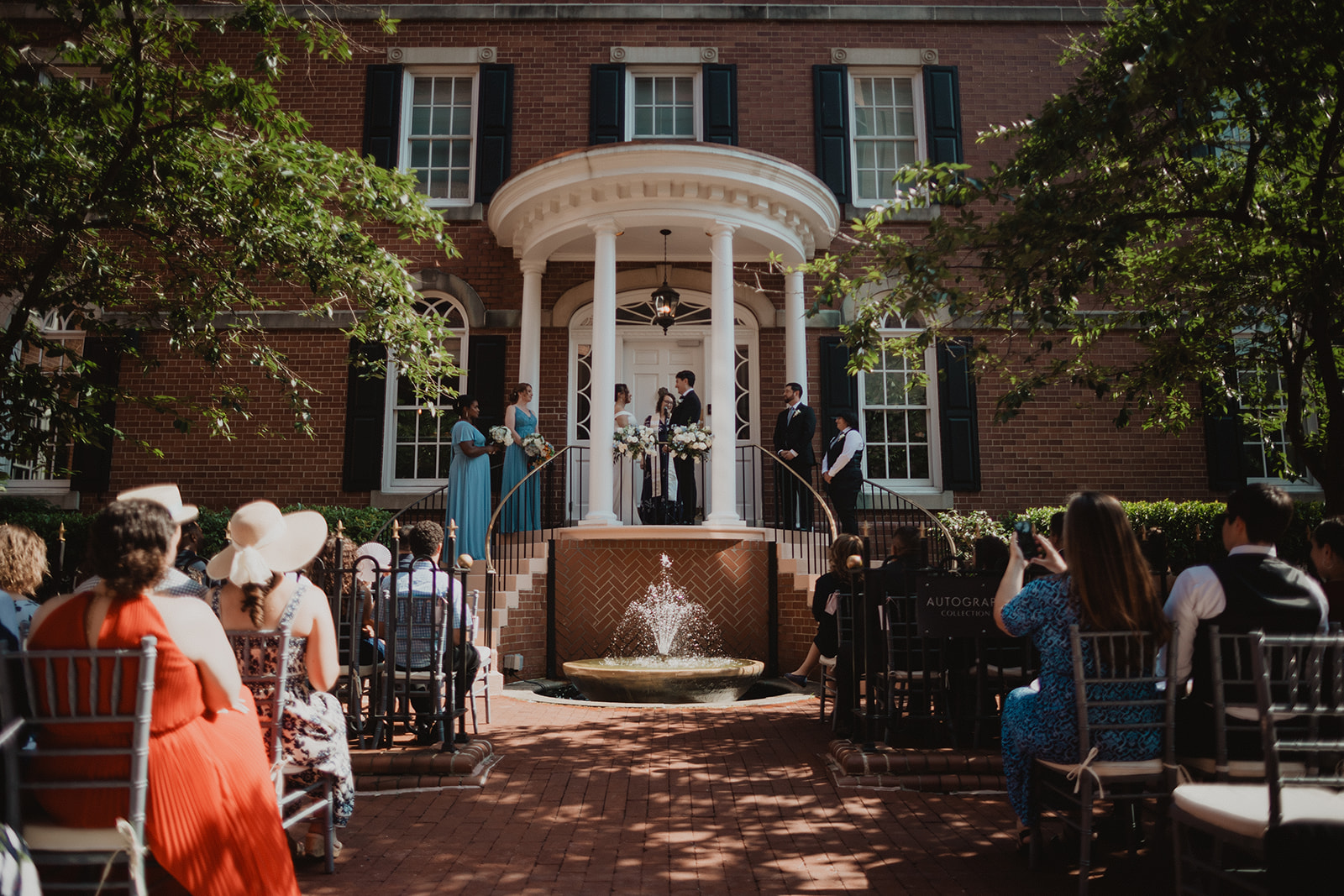 Jessica and Nicholas saying vows on the portico steps of the Morrison House above the fountain, guests watching from the shaded brick courtyard
