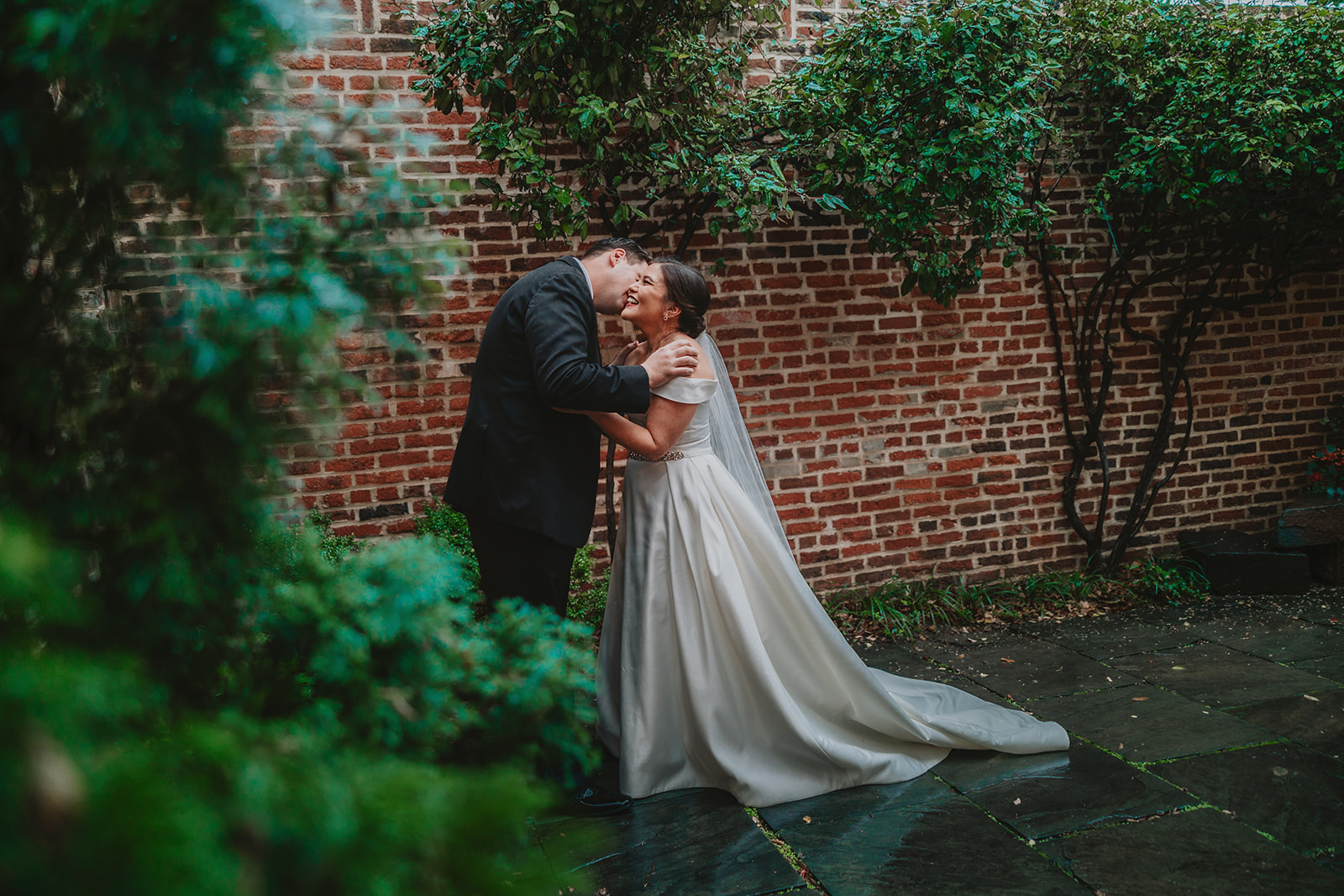 Anthony kissing Marrissa in the walled brick garden at The Athenaeum, shot through deep green foliage