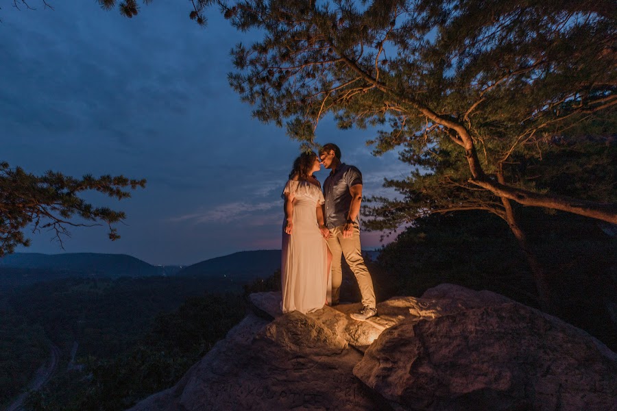 Ashley and Alan lit warm on the rocks at Weaverton Cliffs at night, framed by pine branches against a deep blue sky