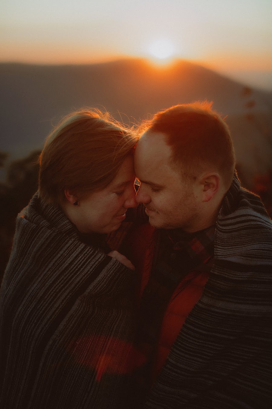 Lilly and Arthur forehead to forehead under a blanket as the sun crests the ridge exactly between them