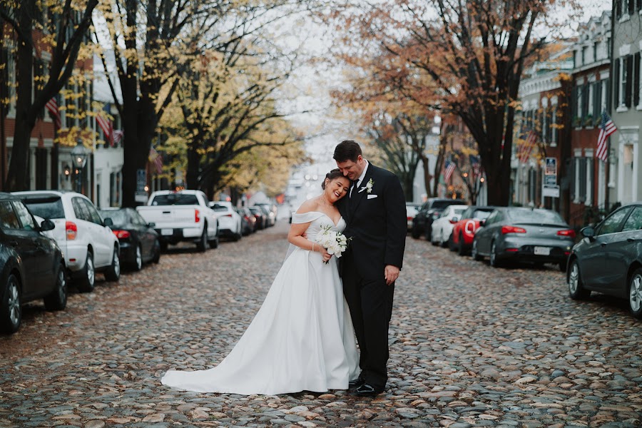 Marrissa laughing into Anthony's shoulder in the middle of a cobblestone street in Old Town Alexandria, flags and autumn trees down both rows of houses