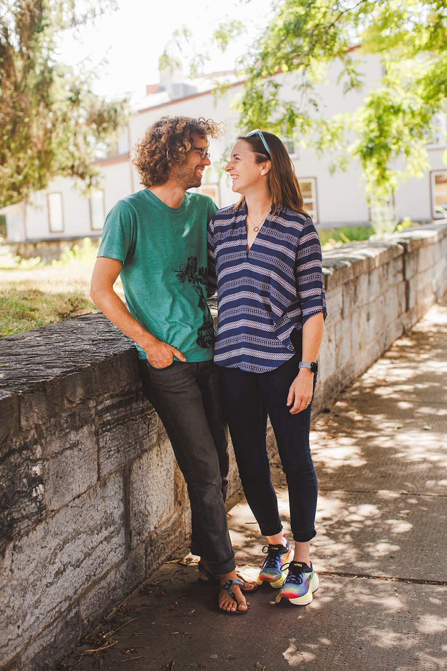 Meredith and John laughing nose to nose against a low stone wall in downtown Shepherdstown, dappled summer light on the sidewalk