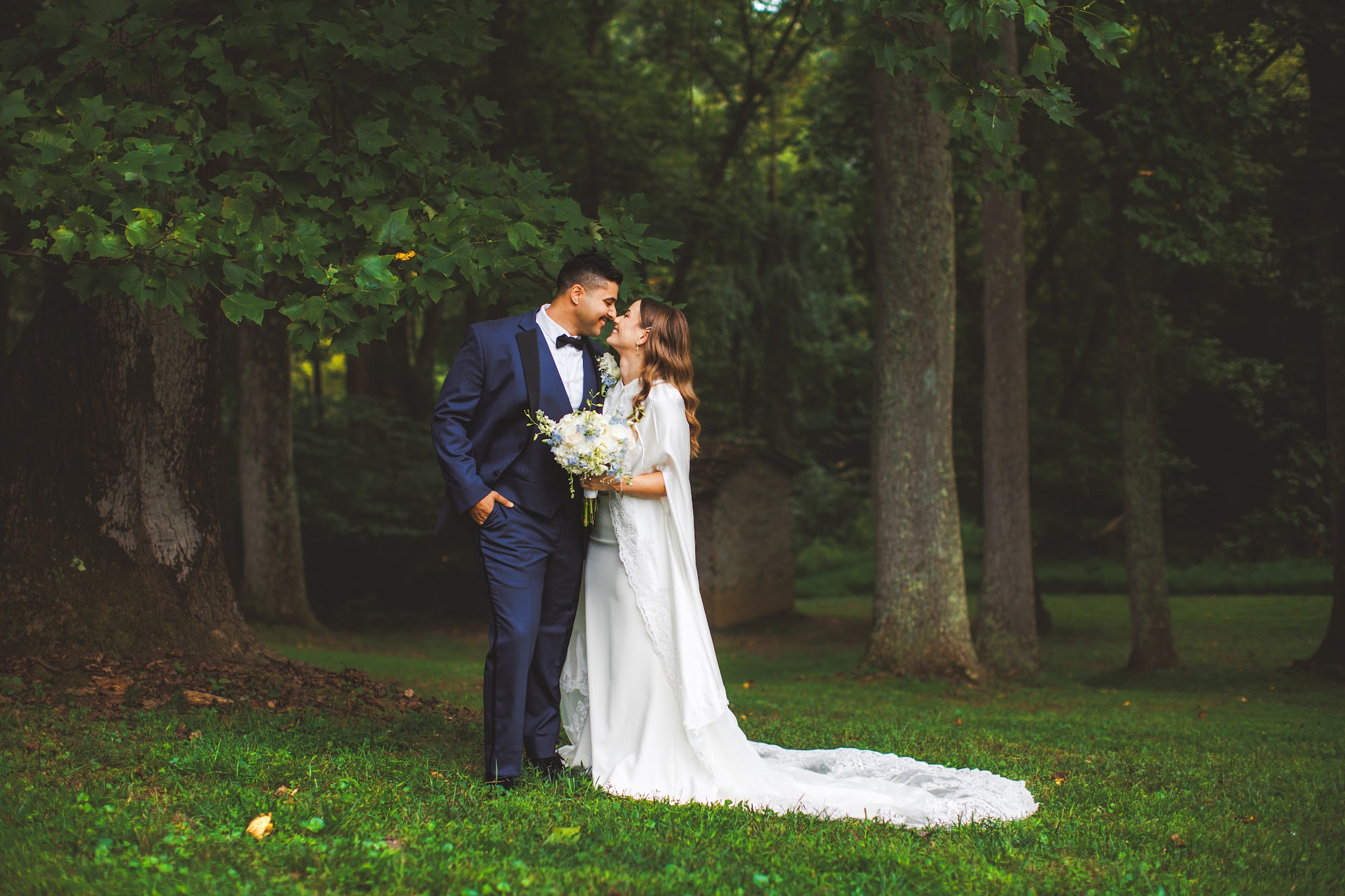 Bailey and Youssef nose to nose beneath tall trees, her lace train spread across the grass