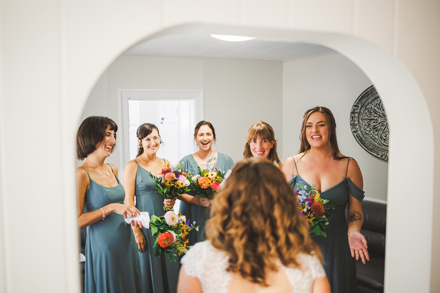 Bridesmaids in sage-green dresses laughing and beaming at the bride during a first look in a bright room