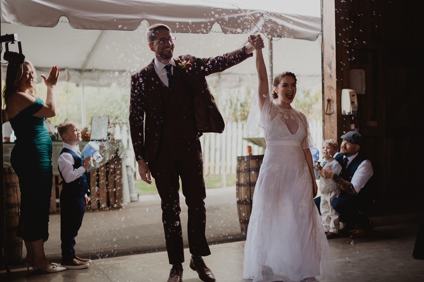 A groom in a maroon suit lifting the bride's hand in triumph as they enter their reception while guests blow bubbles