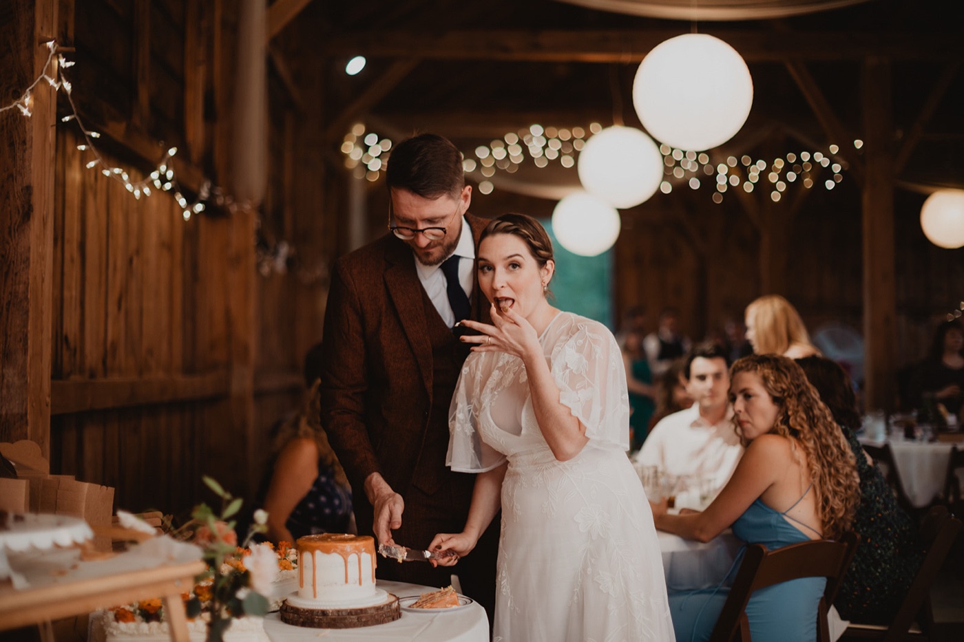 A bride tasting cake off her finger with a playful look as the groom slices their drip cake at a lantern-lit barn reception