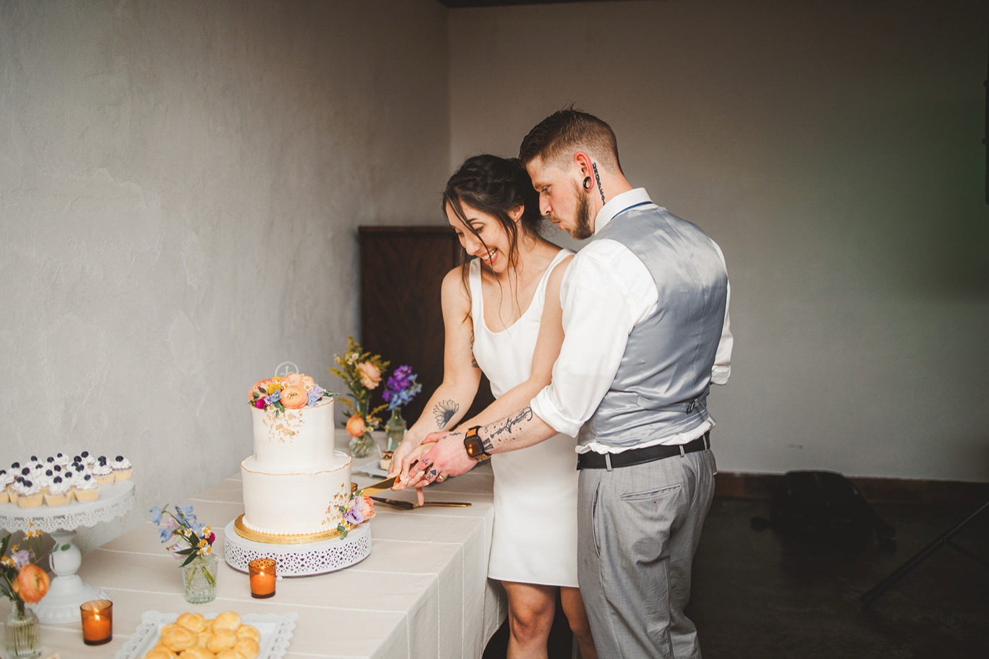 A groom standing behind the bride and guiding her hands as they cut their two-tier cake beside a dessert table