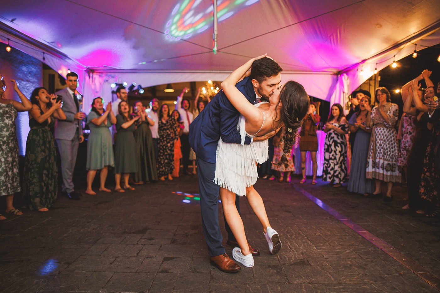 A groom dipping and kissing the bride on the dance floor under colorful lights as guests circle around clapping