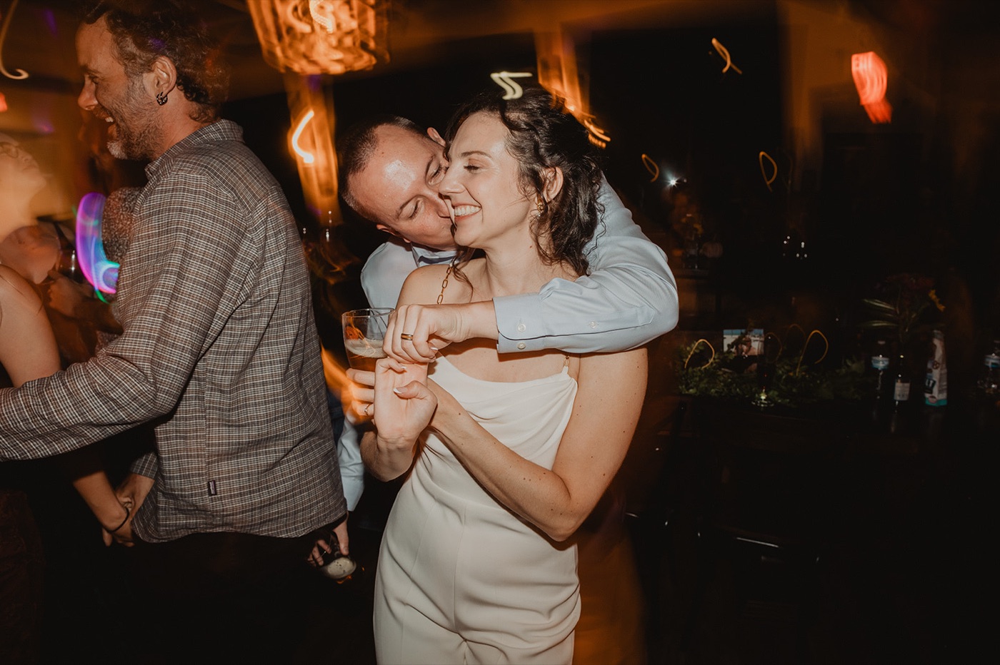 A bride laughing with her eyes closed as the groom nuzzles her cheek from behind on a dim dance floor