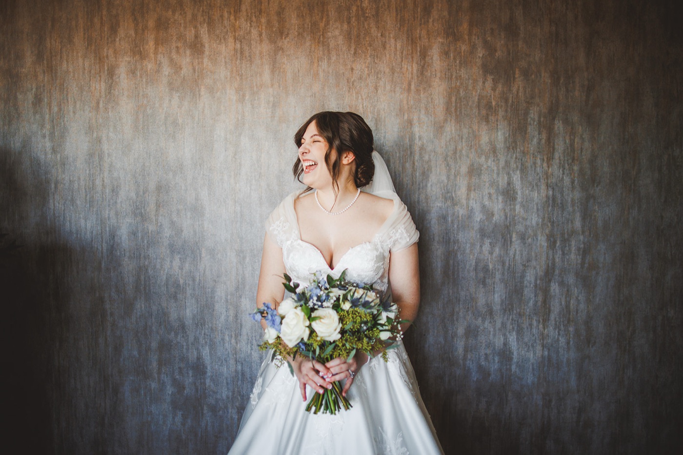 A bride in a lace ballgown and veil laughing with her head turned, holding a white-rose and blue bouquet against a rust-toned wall