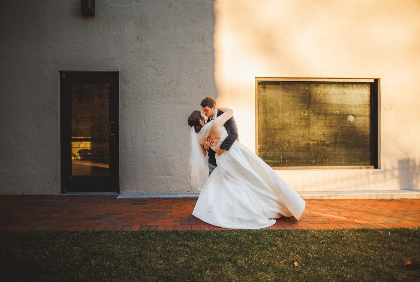 A groom dipping the bride into a kiss on a brick patio beside a stucco building in golden evening light