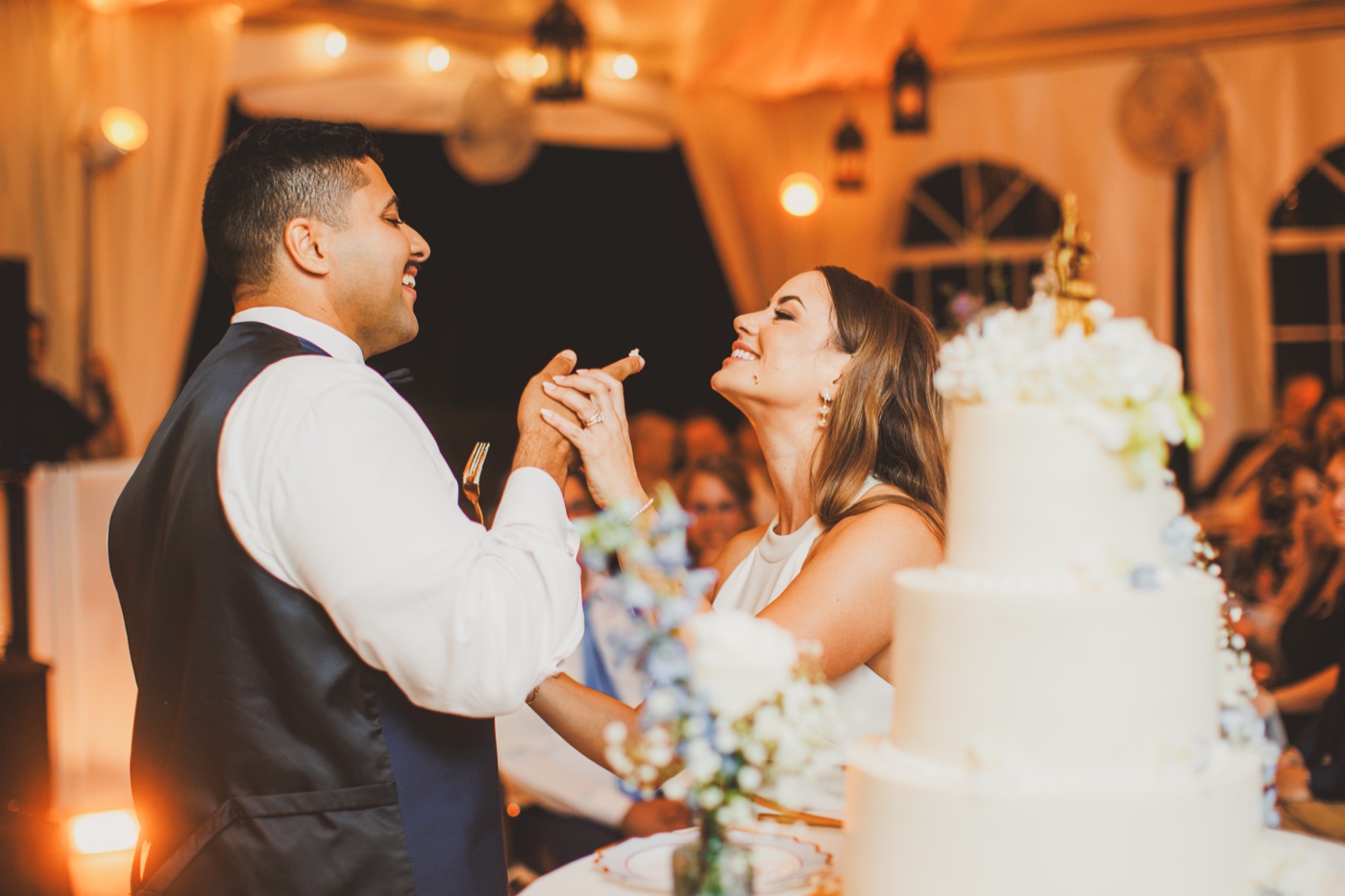 A bride and groom holding hands and smiling at each other beside a tall tiered cake in a warmly lit tented reception