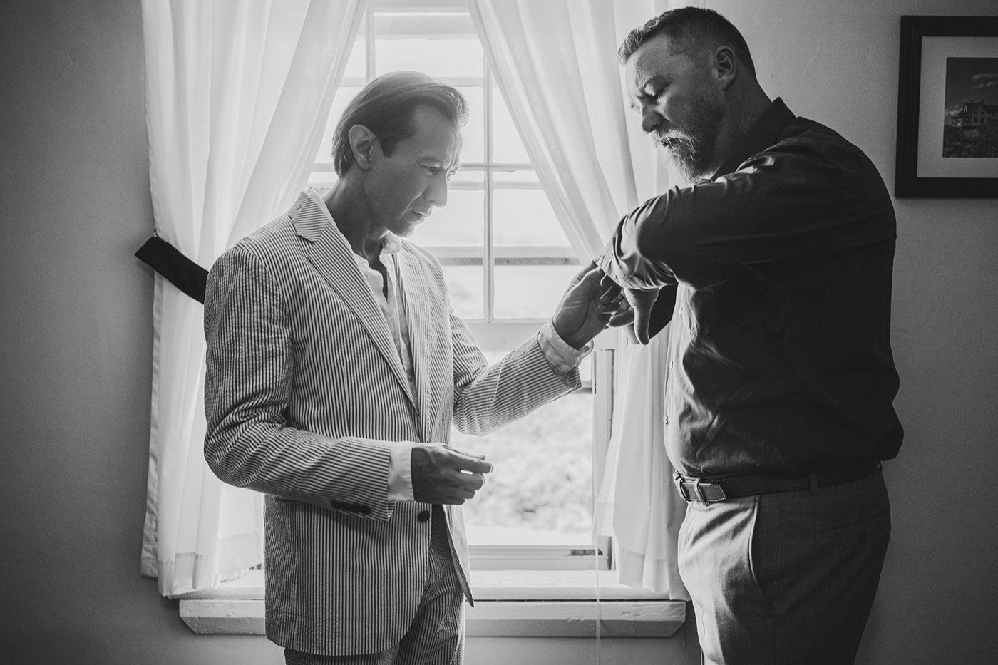 A black and white frame of one man in a seersucker suit helping another fasten his shirt cuff by a curtained window